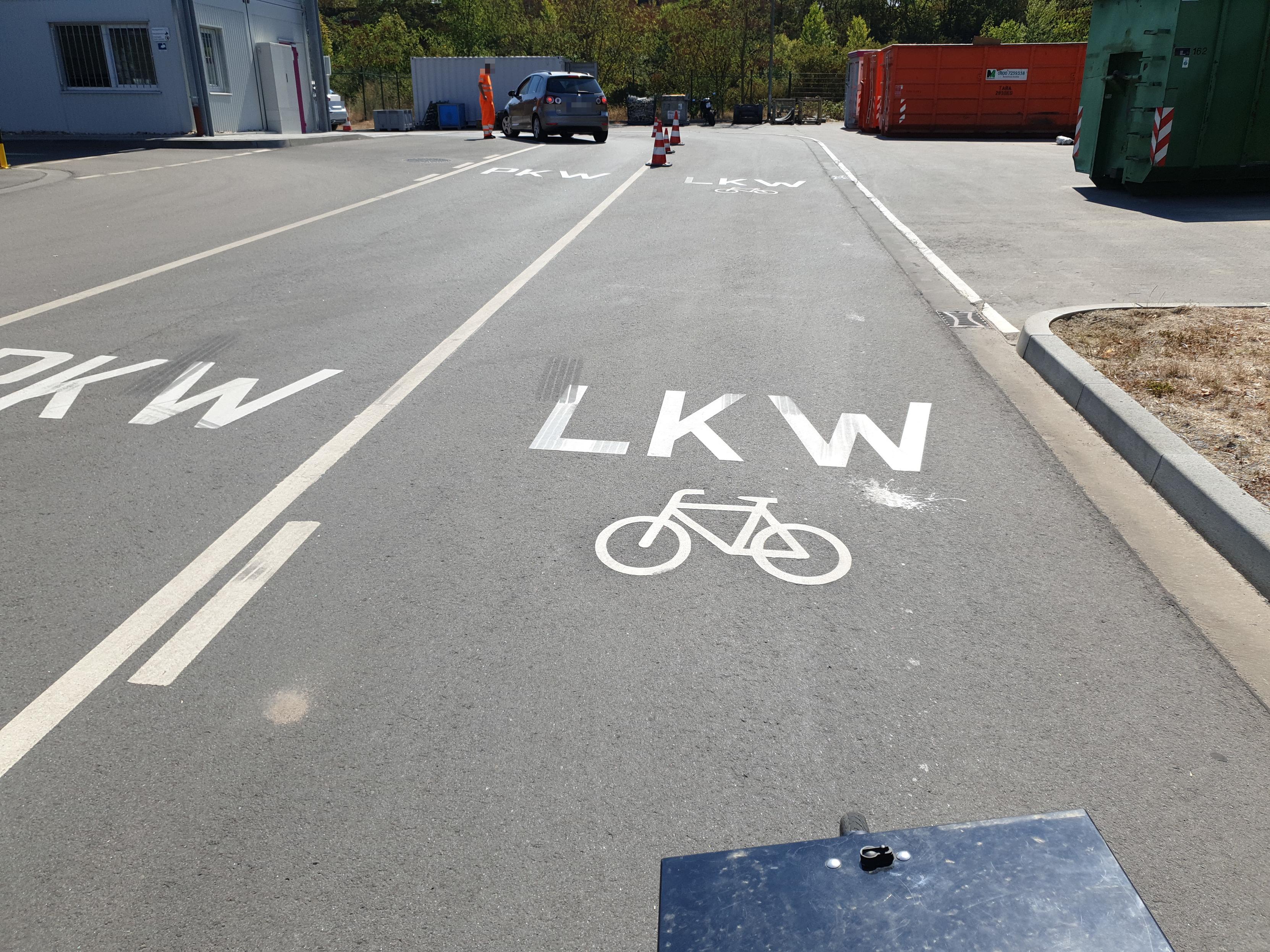 Die Einfahrt zu einem Recyclinghof mt zwei Fahrstreifen. Der linke ist mit einer Markierung „PKW“ versehen, der rechte mit einer Markierung „LKW“ und darunter einem Fahrrad-Piktogramm. Unten am Bildrand ist noch die Front eines Lastenfahrrads mit einer schwarzen Alukiste zu sehen. Im Hintergrund steht eine Person im orangefarbenen Anzug neben einem PKW und spricht mit den Insassen.