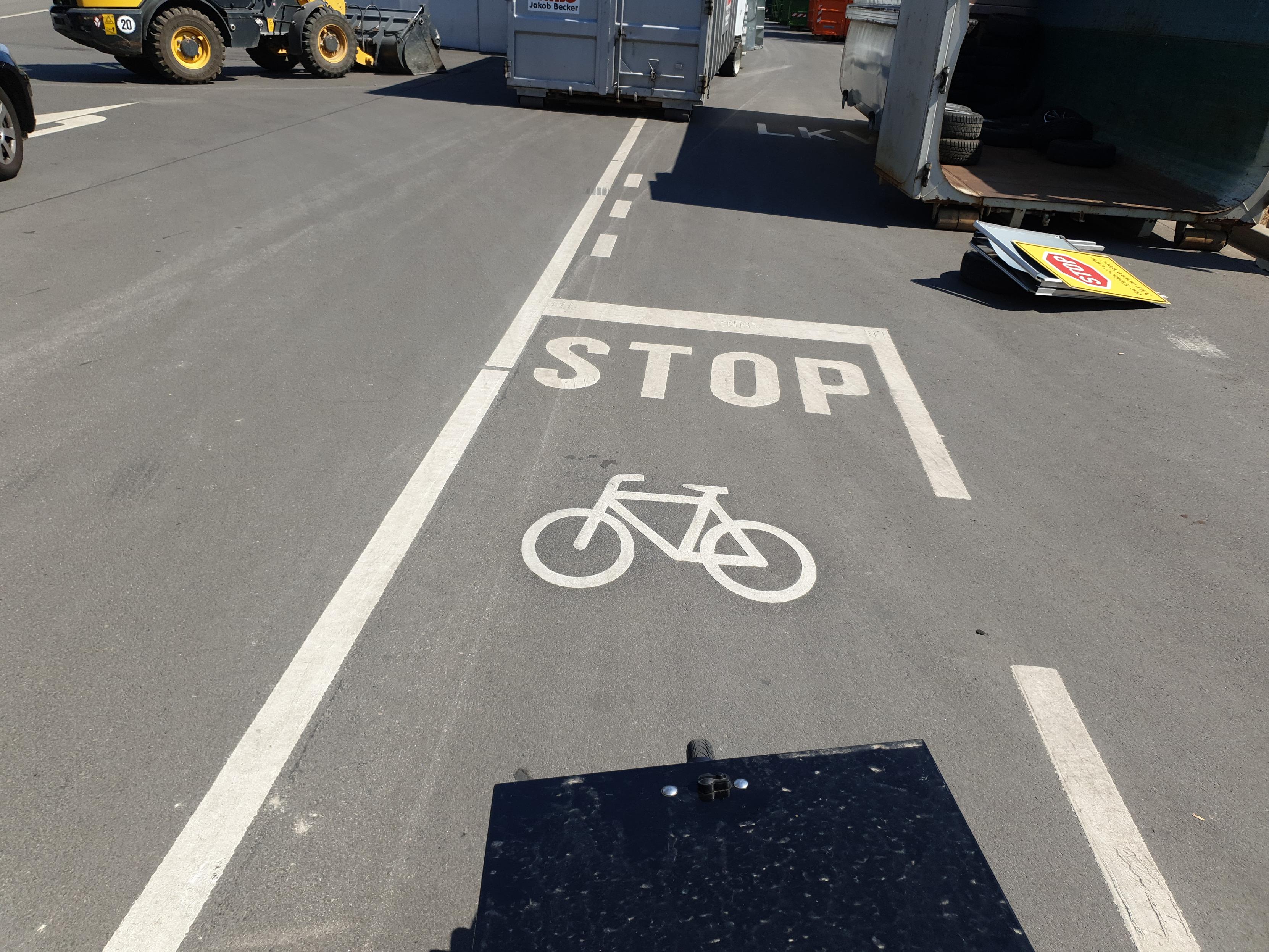 Markierung eines Radfahrstreifens auf einem Recyclinghof mit einer Markierung „STOP“ und einem Fahrrad-Piktogramm darunter. Am unteren Bildrand ist wieder die schwarze Kiste eines Lastenfahrrads zu sehen. Im Hintergrund ein Radlader und diverse Container.
