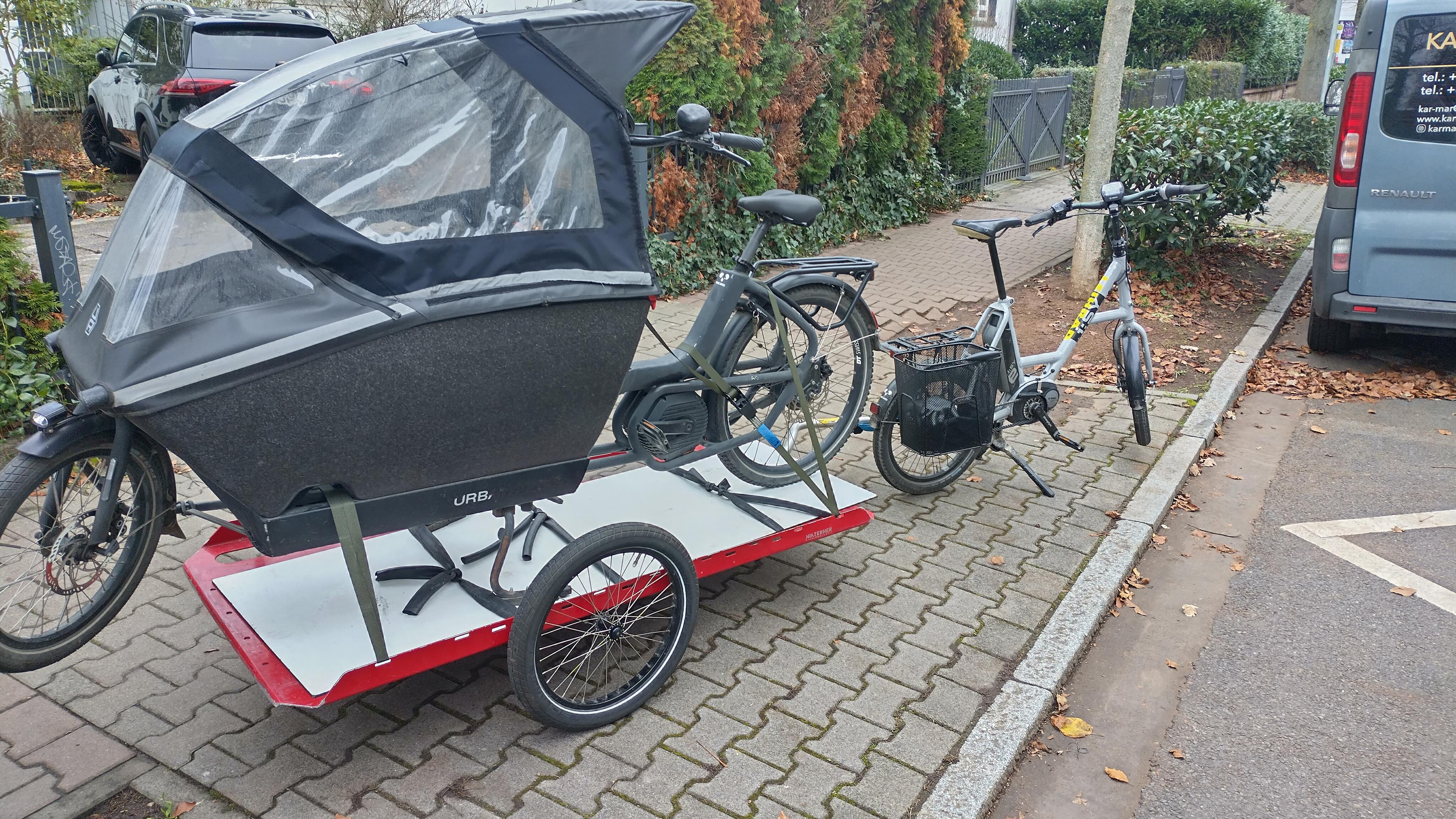 A small, grey, 20 inch compact bike towing a long red and white platform trailer. There is a big Urban Arrow cargo bike strapped onto the trailer.